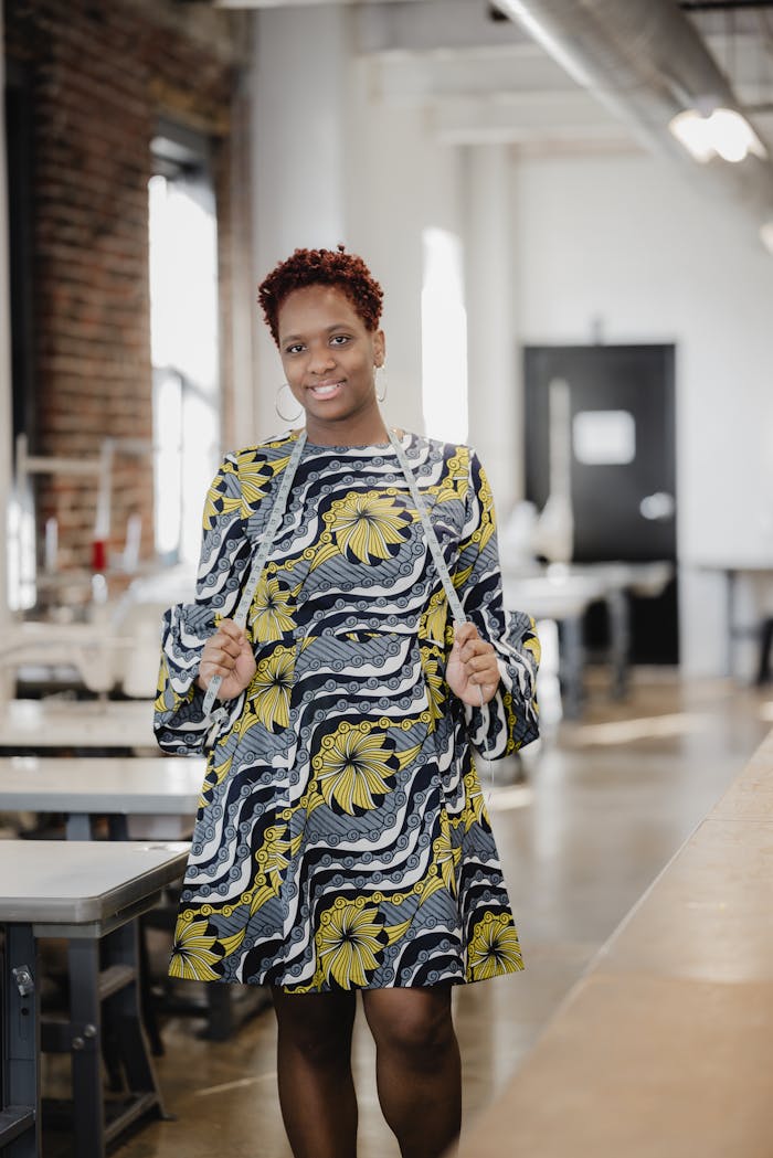 A confident woman smiling in a patterned dress with a measuring tape in a workshop setting.